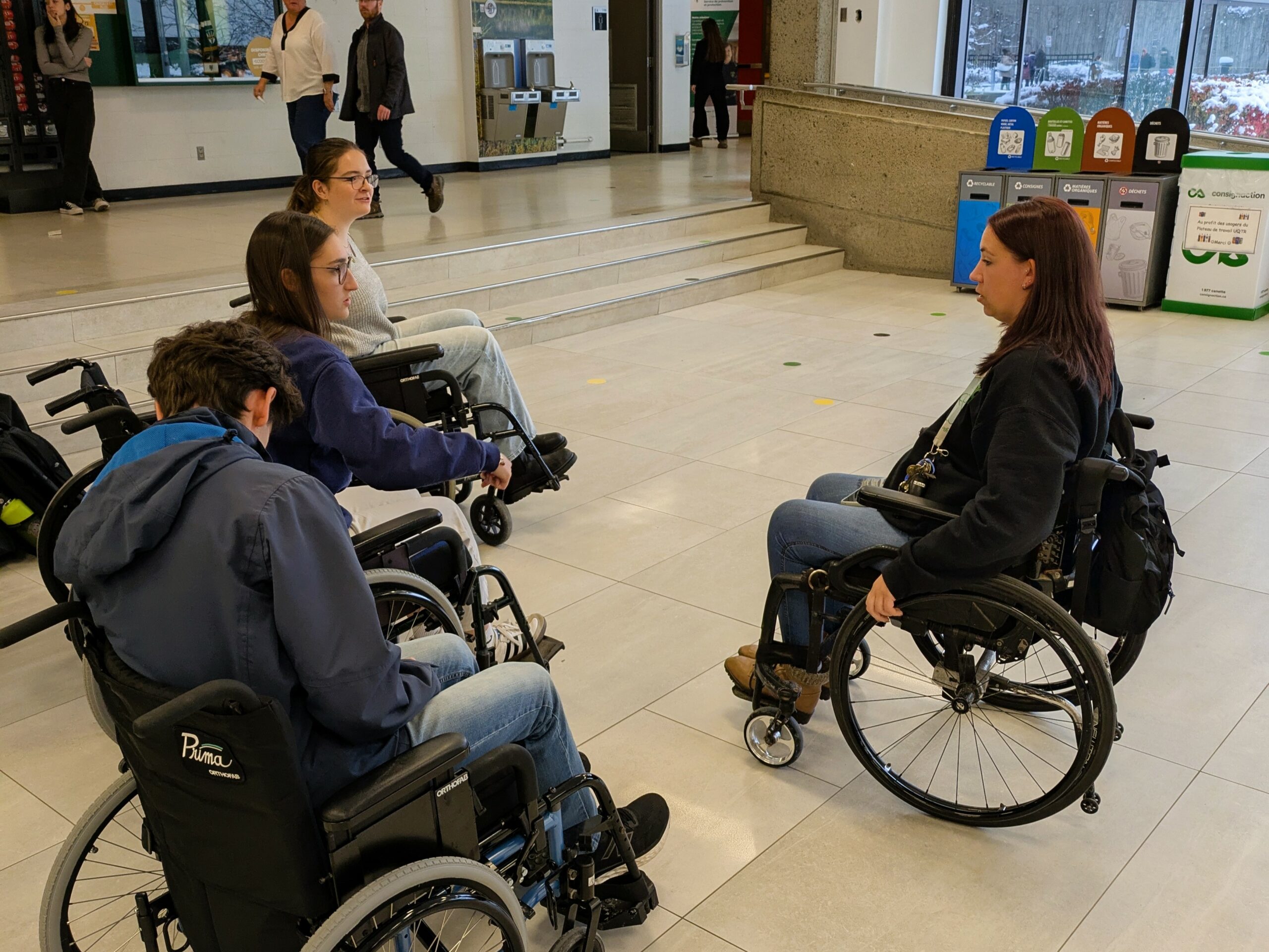Un petit groupe de personnes étudiantes teste des fauteuils roulants sous la supervision de Chloé, une étudiante en situation de handicap moteur et organisatrice de l'événement.