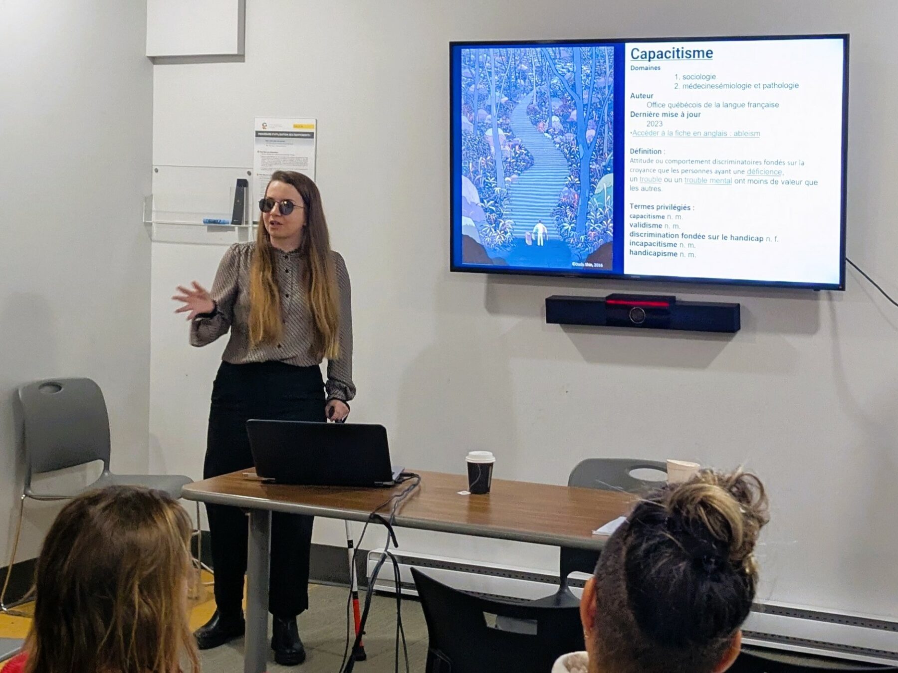 Maude Chartier-Desjardins, une femme blanche en situation de handicap visuel, présente une diapositive sur le capacitisme devant un groupe. Elle porte des lunettes de soleil et s'appuie sur sa canne blanche.